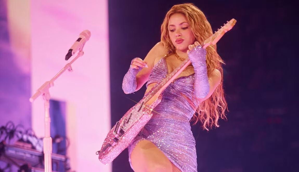 Artista tocando una guitarra eléctrica brillante durante un concierto en vivo con iluminación rosa y vestuario brillante.