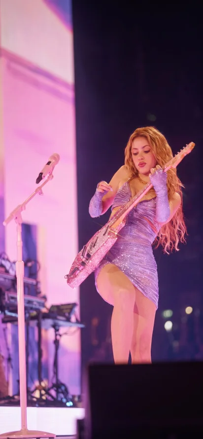 Shakira tocando guitarra eléctrica en concierto con vestido brillante morado.